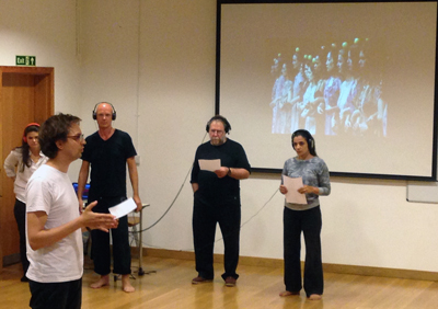 Participants during the Chorus in Action Workshop: 4 adults with headphones on, 1 man in foreground speaking, in front of a screen with a video projection on it