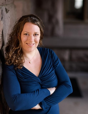 A photo of Rachel Bryant Davies, leaning against a brick wall with her arms crossed, smiling.