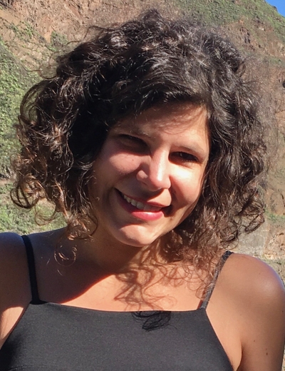 A photo of Giovanna, smiling, standing in front of a mountain in the sunshine.