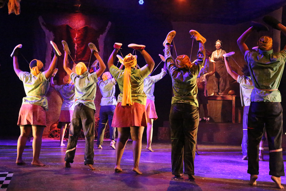 Dress rehearsal photograph of the 2022 play iKrele leChiza, depicing a group of performers, from behind, stood barefoot with their arms raised and shoes on their hands