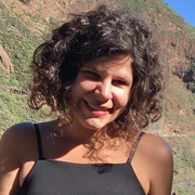 A photo of Giovanna, smiling, standing in front of a mountain in the sunshine.