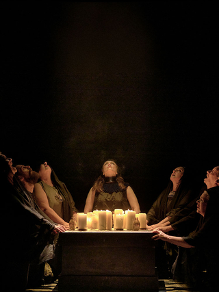 A photograph from a performance of Na Peirsigh, showing actors gathered around a table full of candles, looking upwards.