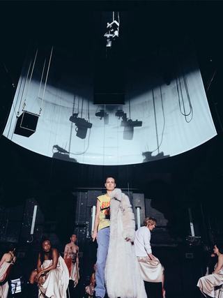 Production photograph of Brie Larson centre stage as Elektra, with a floor-length fur coat slung over one should and a microphone in the other hand, she has a shaved head and is wearing a yellow punk-style t-shirt.