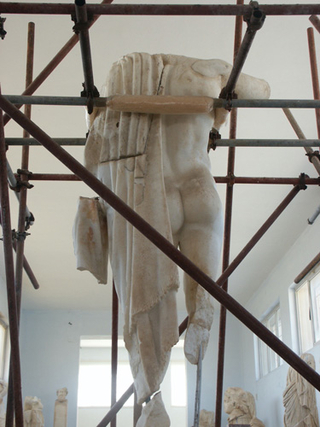 Photograph of an ancient Greek statue, without a head, surrounded by scaffolding